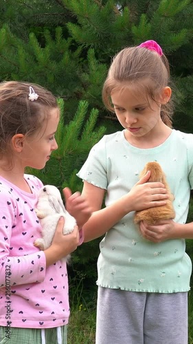 Happy girls hold and pet little baby rabbits outdoors in garden. Children care pets. Kids play with animals. Agriculture, farming. Vertical shot