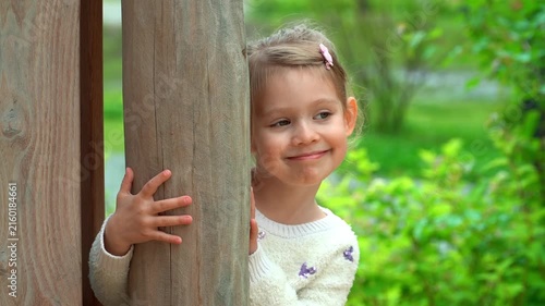 Portrait of little girl hides on playground and laughs. Happy child plays in green summer park. Kid leisure outdoors.