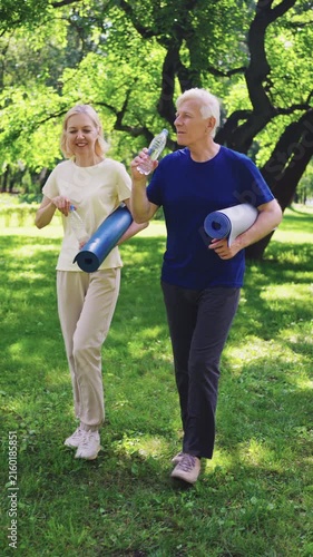 Active senior couple walking on green grass in sunny park. Mature man and woman carrying yoga mats and drinking water after outdoor workout