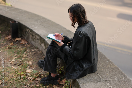 Young woman sits on stone wall beside quiet road, writing notes in small notebook with blue pen. She wears black jacket, dark jeans, and chunky shoes, showing quiet focus in soft natural light