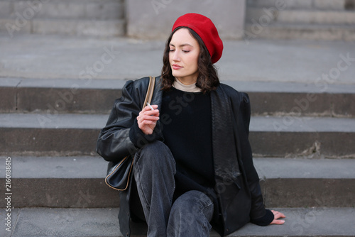 Young woman rests on concrete steps in black coat, dark sweater, and dark jeans, holding cigarette with calm pensive mood in muted urban setting, showing relaxed street fashion style