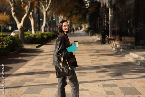 Young woman in dark outfit stands on sunlit urban sidewalk holding coffee cup, turquoise notebook, and handbag, looking toward camera with calm expression, autumn trees and softly blurred street