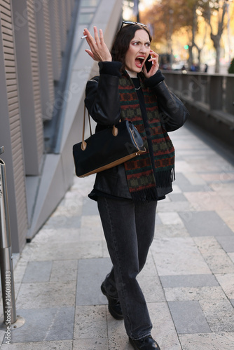Young woman in dark clothing stands on sidewalk during heated phone call, raising hand with cigarette, handbag on arm, soft autumn daylight, city backdrop, intense frustration and conflict