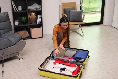 Young woman kneels beside open suitcase while packing folded clothes in bright modern living room, with organized shelves, cushioned chair, light wood floor, and daylight from large window