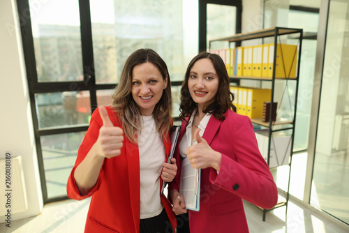 Two confident women smile and give thumbs up in bright modern office, holding folders and documents near large windows, shelves of binders, warm daylight, corporate success atmosphere