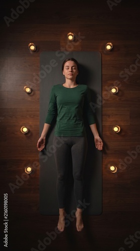 Vertical photo of a peaceful young woman practicing meditation in corpse pose on a yoga mat with candles