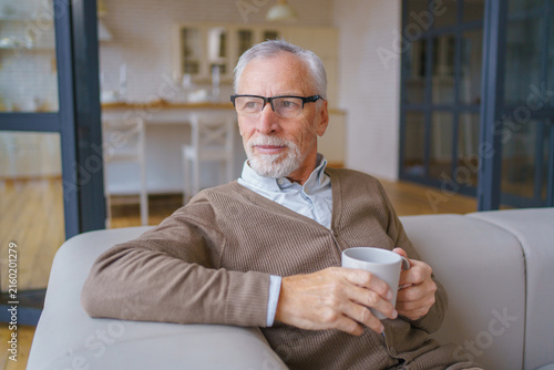 Senior man holding cup and sitting on couch in living room, smiling elderly male looking away at home, relaxed lifestyle, cozy interior and retirement concept