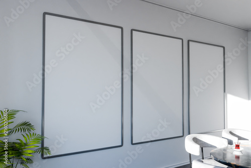 Three blank vertical picture frames hanging on a white wall in a modern living room with a green plant and white upholstered chairs around a round table