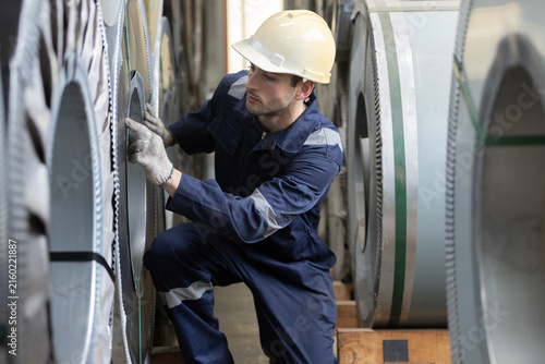 Obraz na płótnie Factory worker inspects steel coils in an industrial warehouse, representing metal manufacturing, quality control, material inspection, workplace safety, production operations
