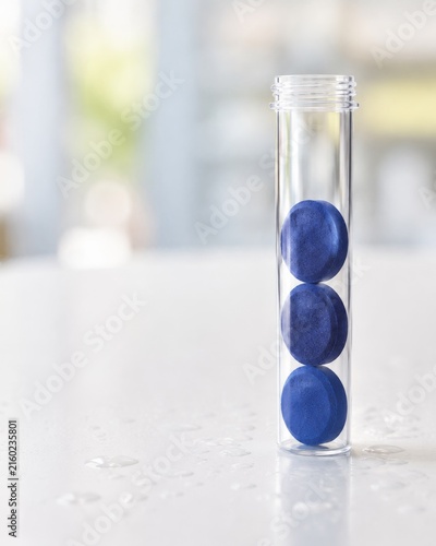 Un tubo transparente contiene tres pastillas azules sobre una mesa de superficie blanca en un espacio iluminado