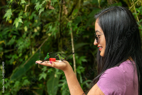 Woman experiencing nature and feeding two vibrant green hummingbirds from her open hand, enjoying an intimate wildlife encounter in a tropical forest setting
