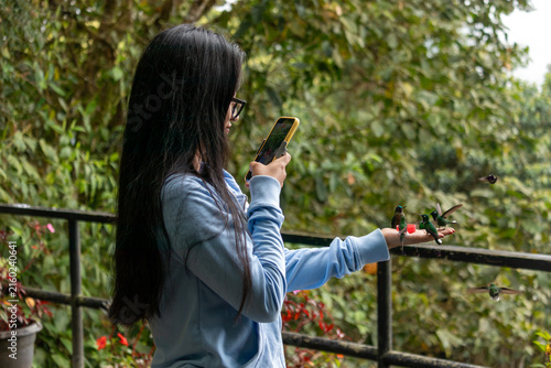 Woman standing outdoors, holding out a hand to hummingbirds feeding from small cups while using a smartphone to record the close encounter with nature and local wildlife