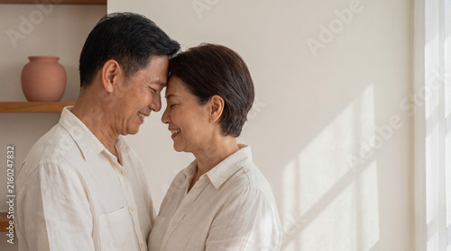 Middle-aged Asian couple smiling and touching foreheads affectionately indoors at home