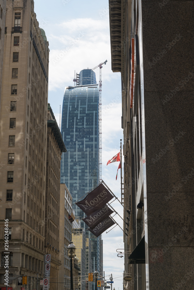 Fototapeta premium narrow view of Yonge St with flag banners outside Moores Clothing for Men, old office towers and (at center) L-Tower condominium and beyond that SkyTower, Toronto
