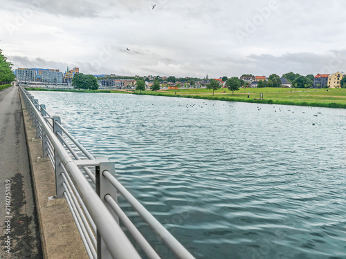Urban riverside walkway with metal railing beside calm water under cloudy sky. Water Mirror lake in Oslo