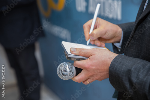 Male reporter writing down key information at official corporate media event briefing. Close-up of journalist hands holding microphone and taking notes with pen on notepad during press conference.
