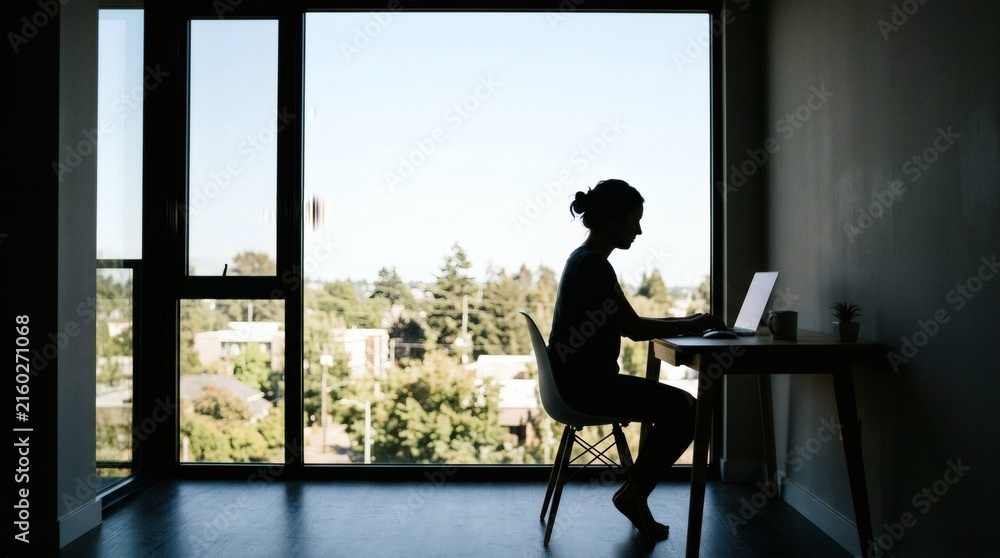 Fototapeta premium Silhouette of woman working on laptop at desk by large window with city view