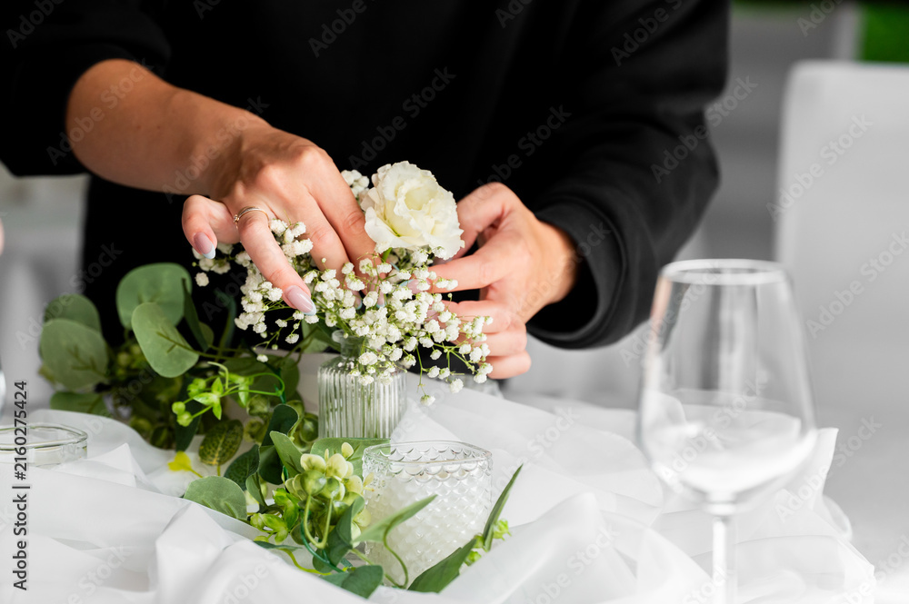Fototapeta premium A woman's hands arranging delicate white baby's breath and a rose in a small glass vase on a decorated wedding table.