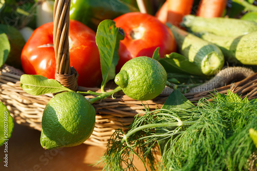 Фотография Fresh harvest display featuring bright green lemons on branch, ripe tomatoes, zucchini, and dill sprigs in a rustic wicker basket