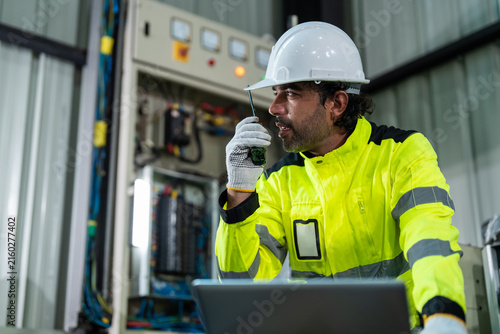 Professional electrical engineer using walkie-talkie and laptop computer for checking power control system in smart factory. Industrial maintenance and automation technology concept.