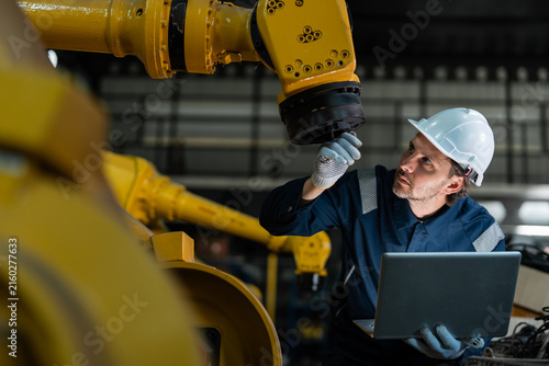 Professional male engineer in a blue uniform and hard hat using a laptop to inspect and program a yellow industrial robotic arm in a modern smart factory, focusing on industry 4.0 automation.