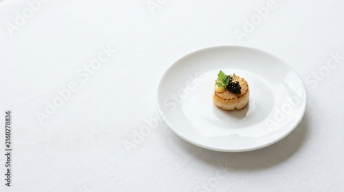 A single seared scallop rests on a white plate. A small dollop of black caviar sits atop the scallop.