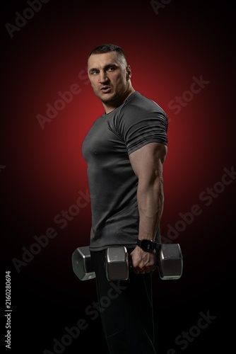 Obraz na plátně Strong athletic male bodybuilder holding heavy steel dumbbells while posing for a portrait in a dark studio setting with a dramatic red circular spotlight background