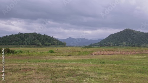 Static wide view of a forested green hill and mountain ridges behind a flat grass plain with a fence under grey storm clouds