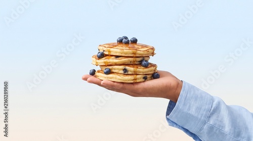 Person holding stacked blueberry pancakes drizzled with syrup against soft pastel blue background with golden brown blueberry