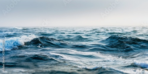Deep blue ocean waves roll across the vast surface during a calm morning with gentle white foam crests under a soft and hazy gray horizon, minimal seascape