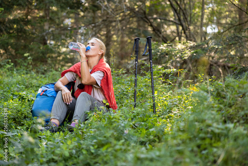 Woman resting from a hike, drinking refreshing water from a plastic bottle while sitting on lush green foliage in a forest
