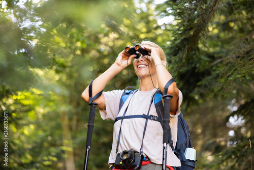 Smiling woman enjoying outdoor adventure, exploring nature with binoculars and trekking poles