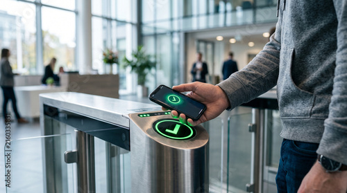 Man using mobile phone for contactless entry at modern stainless steel turnstile gate in office lobby during bright daytime with green verified checkmark status icon
