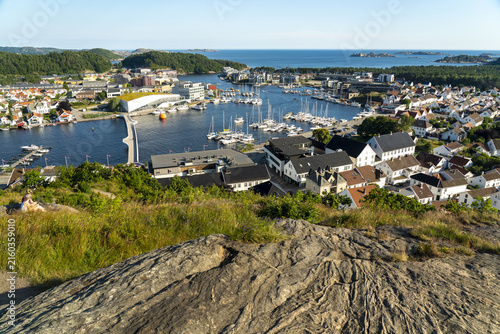 View of the coastal town of Mandal with its harbor, numerous boats, white houses, and a modern building connected by a bridge, seen from a rocky vantage point in Mandal, Agder, Norway.
