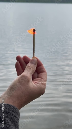 Lit match burning steadily above calm lake water. Flame flickering in hand against a peaceful natural backdrop. Concept scene showing fire persisting near water outdoors..