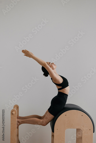 Side profile view of woman in black sportswear arching backward over wooden pilates barrel, arms raised overhead stretching spine and shoulders, kneeling barefoot on padded bench in white studio room