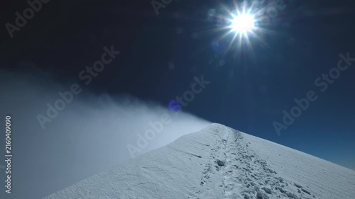 POV handheld perspective of final climber steps reaching Mont Blanc summit 4810m, strong gale wind blowing snow off narrow icy ridge under bright sun, epic mountaineering achievement in French Alps