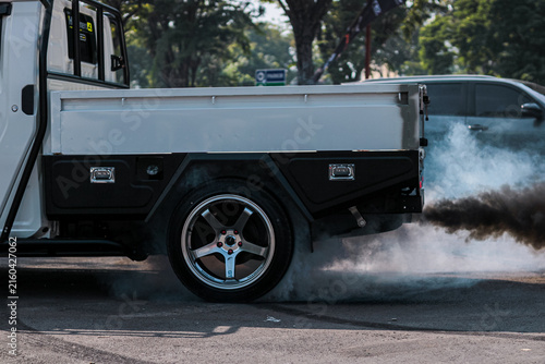 Close-up of a modified white pickup truck performing a heavy tire burnout. Custom flatbed and alloy wheel spinning with thick smoke on the asphalt. Motorsport and automotive tuning concept.