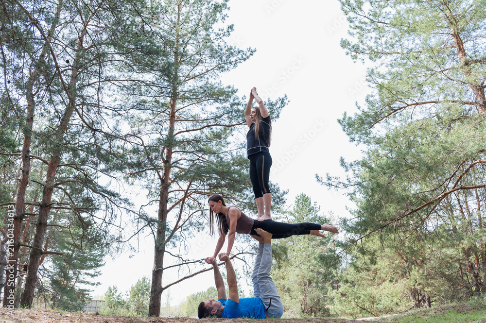 Fototapeta premium Man supports two women performing a challenging three person acro yoga tower with plank and standing balance outdoors in a sunny pine forest.