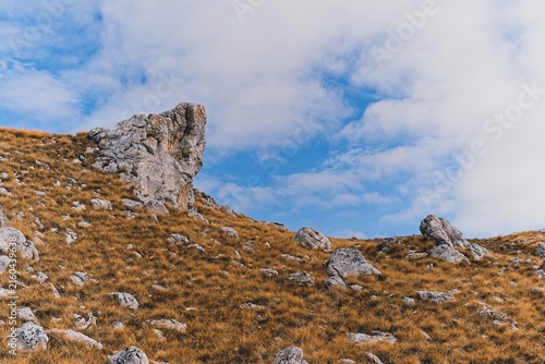 Fotografie A large jagged rock formation standing on a dry grassy hill slope against a clou