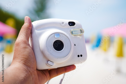 Canvastavla White instant camera held in a person's hand outdoors, with a colorful blurred background, illustrating travel photography, summer vacation, leisure, creativity, and preserving special memories