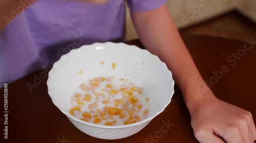 Person in purple shirt holds white bowl of golden cereal with milk while eating breakfast using spoon at table.