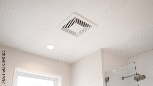 Bright modern bathroom ceiling with a square ventilation grille, recessed light, window light, and part of a glass shower and tiled wall