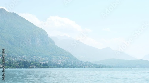 People enjoying water sports, kayaking and paddleboarding on calm surface of Lake Annecy in France, scenic alpine mountain landscape and village settlement on shore during warm august vacation