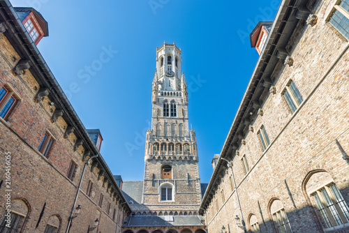 The Belfry of Bruges, a medieval bell tower in Bruges town in Belgium, Europe. Low Angle View of an Ancient Stone Tower Amidst Historic Brick Buildings Under a Clear Blue Sky on a Sunny Day