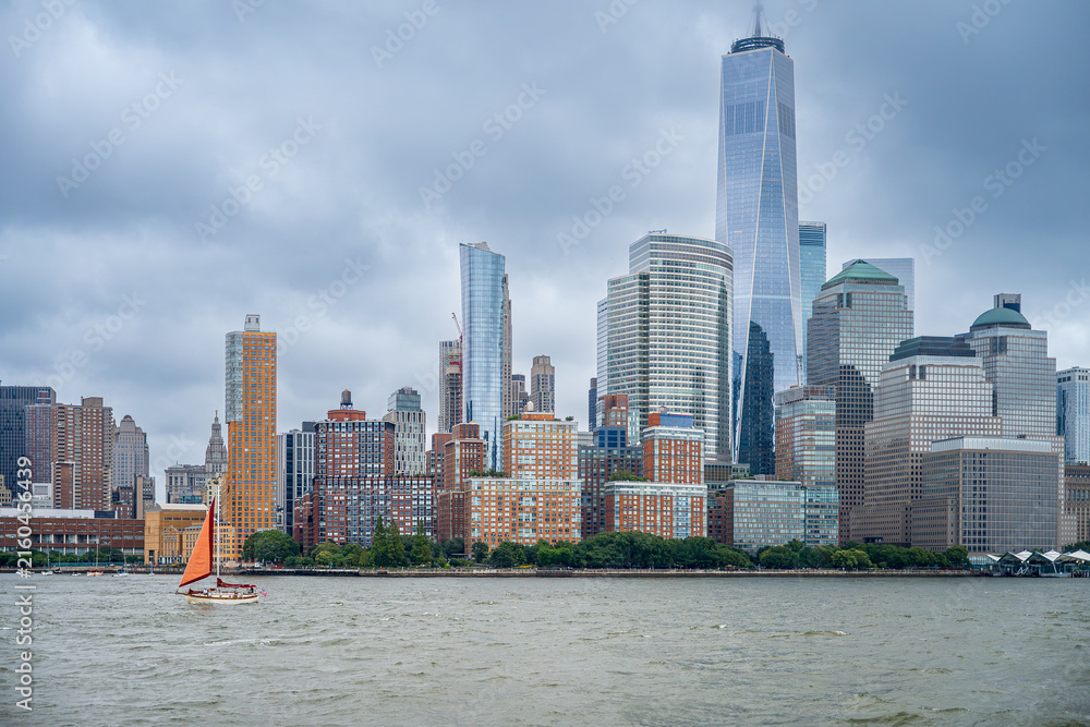Obraz premium Sailboat passing Lower Manhattan and One World Trade Center. Wide river view shows an orange sail beneath clustered towers and a gray overcast sky.