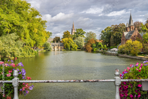 Bruges town in Belgium, Europe. Historic European city lake with lush green trees, blooming flowers, and traditional ancient spires under a dramatic sky