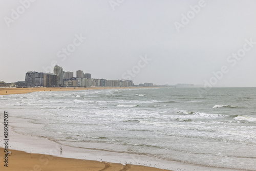 Panoramic view of a grey urban coastline on a cloudy day, featuring ocean waves gently breaking on a wide sandy beach. Ostend town in Belgium, Europe.