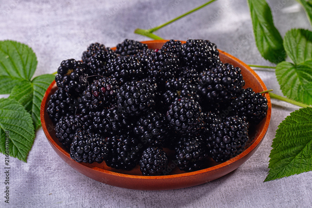 Fototapeta premium Fresh blackberries in wooden bowl with green leaves on gray background