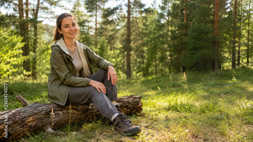 Smiling woman in hiking gear sits on a log in a sunlit pine forest, resting and enjoying nature during a peaceful outdoor hike